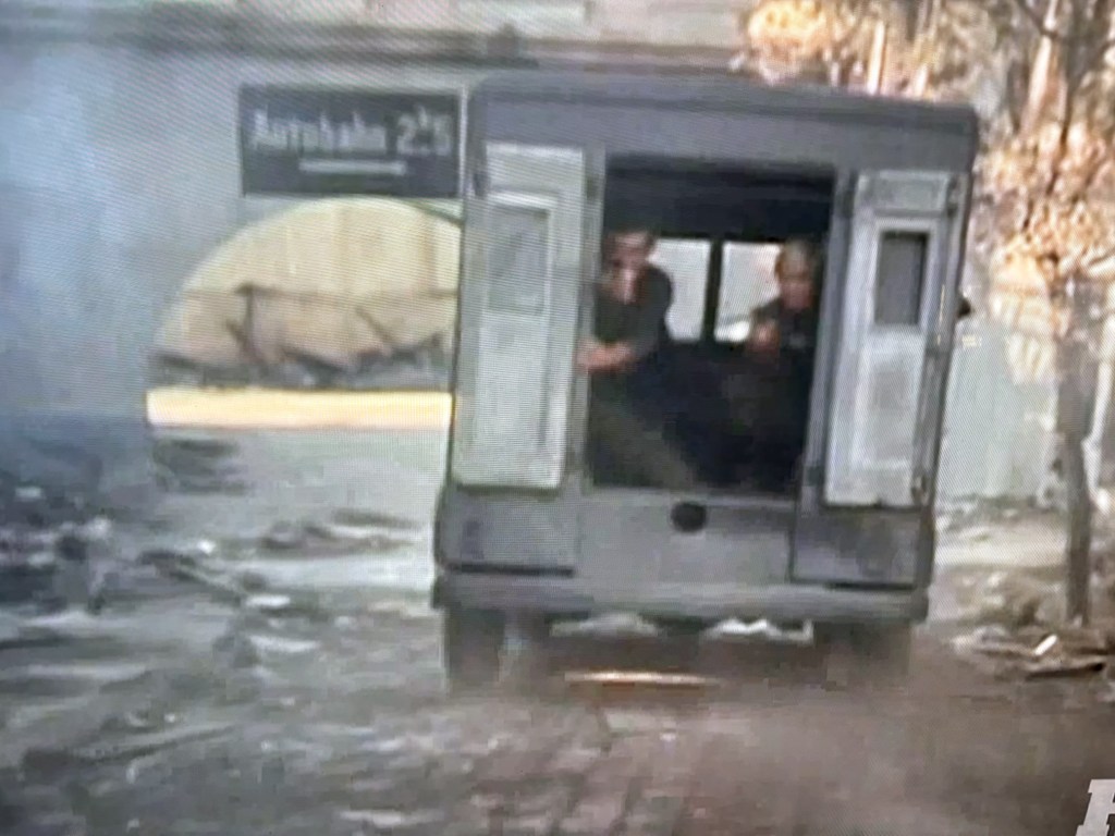 A gray vehicle with open doors, driving down a street with visible debris. Two individuals are seated inside, looking out. A sign above indicates a location.