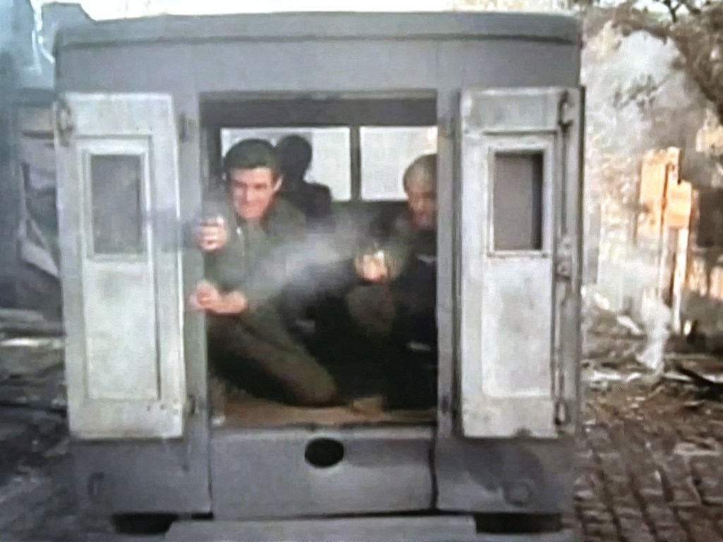 Two men in military uniforms crouch inside a small, damaged structure, firing weapons in a tense situation, with smoke and debris surrounding them.