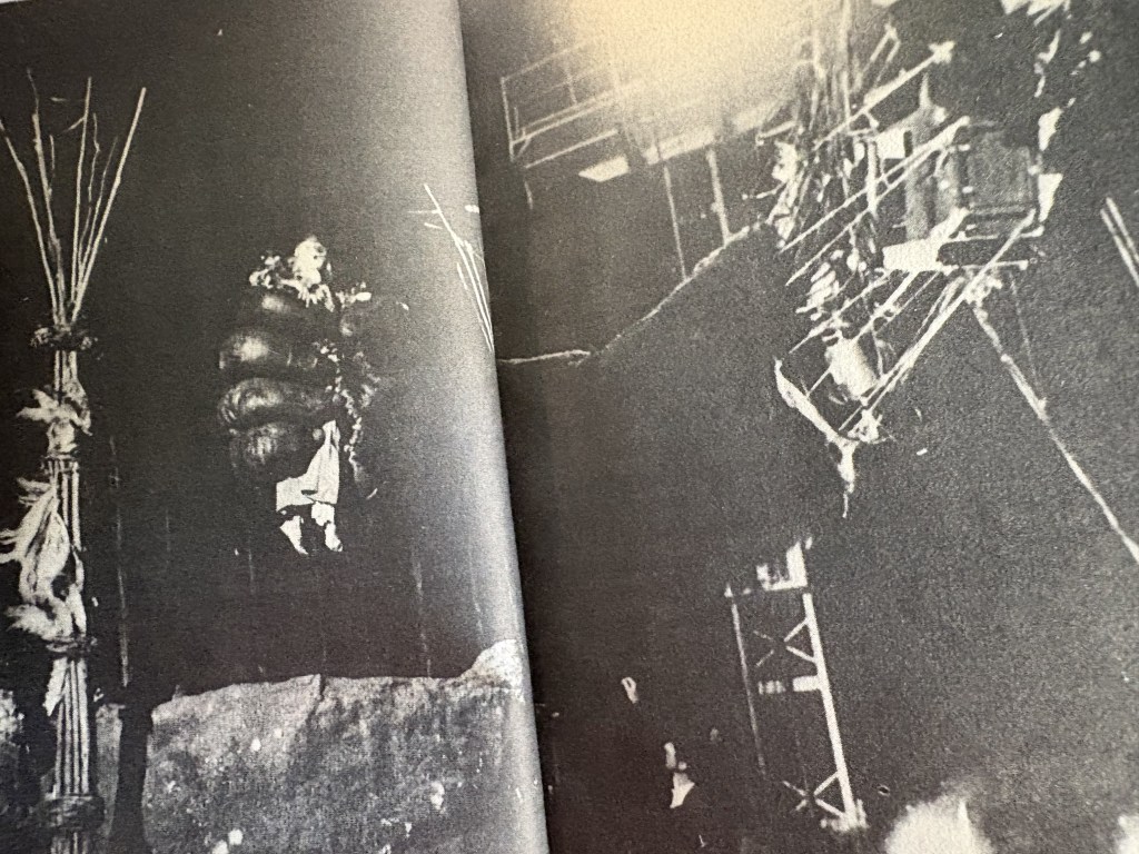 A black and white photograph showing a person suspended in the air, surrounded by large objects that resemble inflatable gear. The background includes industrial structures and rigging.