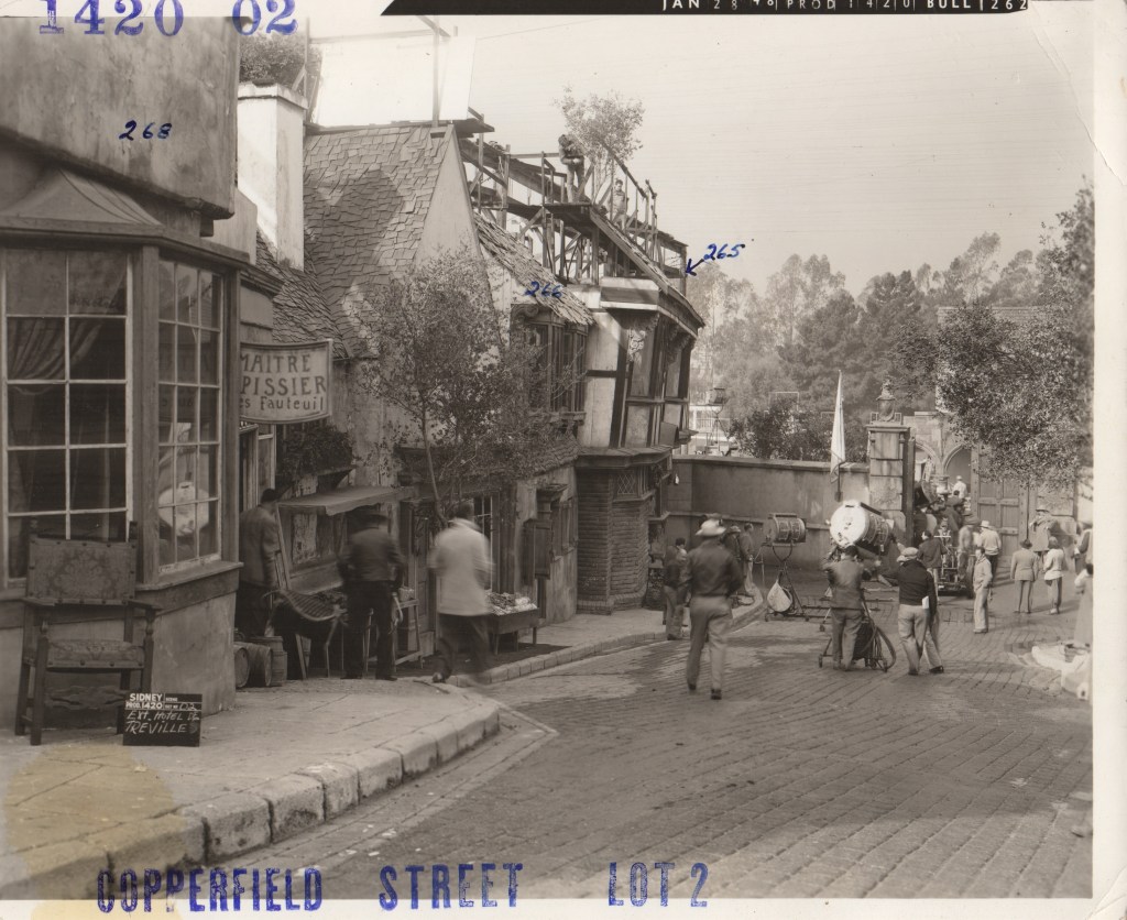 A historic street scene featuring vintage buildings, patrons walking, and crew members setting up film equipment. The setting is bustling with activity, showcasing a mix of architecture and people engaged in various tasks.