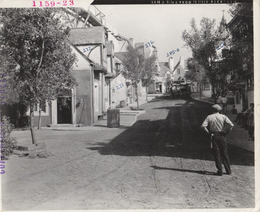 Black and white image of a quiet, sloped street lined with trees and vintage buildings. A figure in uniform stands with their back to the camera, observing the street. Small groups of people are scattered along the sidewalk.
