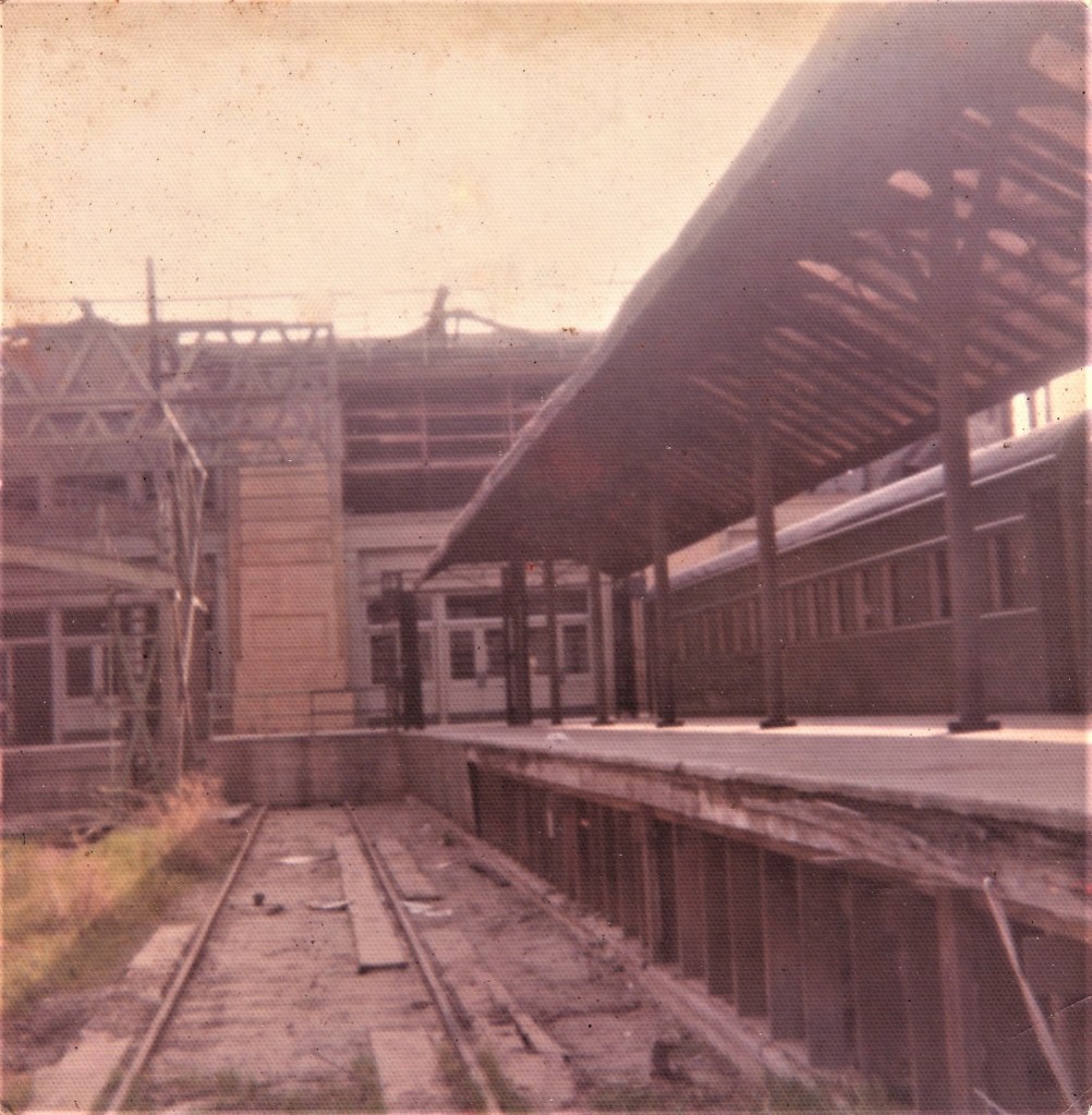 An old railway station with a worn platform, partially covered roof, and rusty train tracks. Distant train cars are visible.