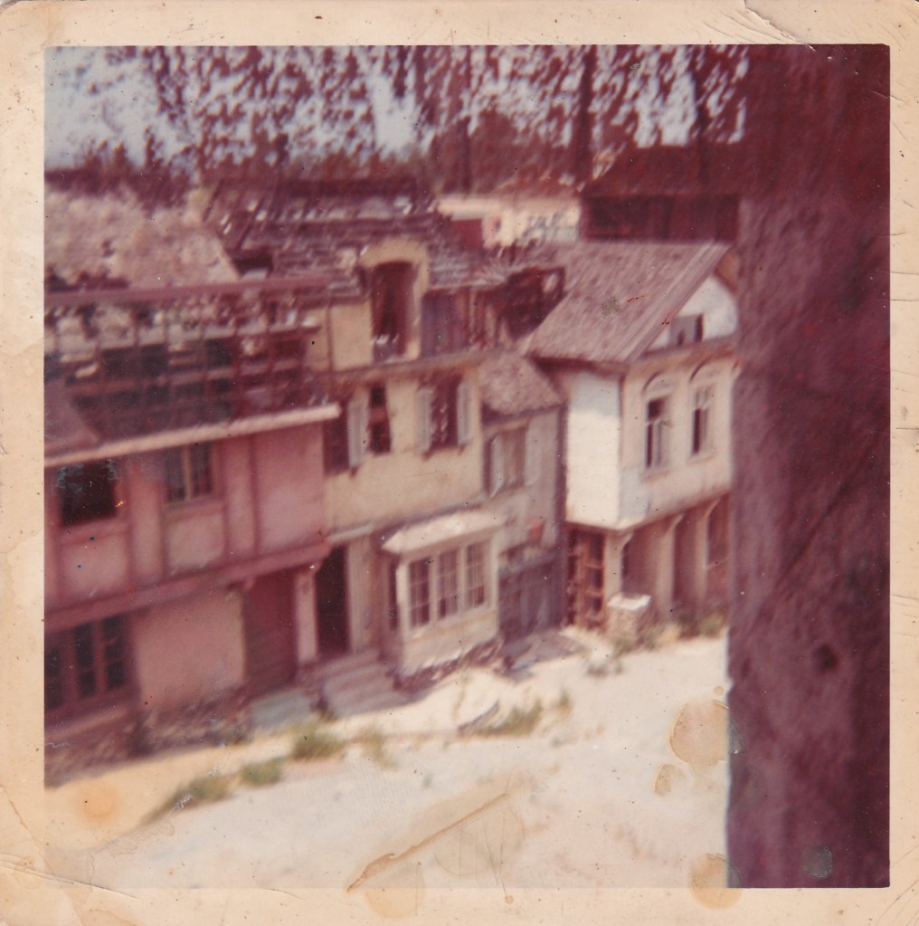 A faded photograph showing a row of old, deteriorating buildings with broken windows and roofs. The scene appears abandoned, with some vegetation visible in the foreground.