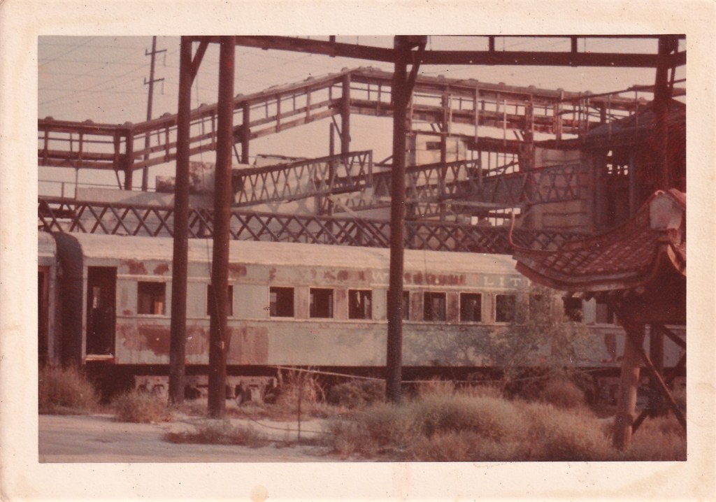 An abandoned train car partially covered by vegetation, surrounded by industrial structures and utility poles.