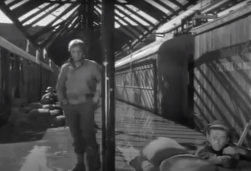 A black and white scene depicting a long corridor with wooden beams, where a man stands casually while another person is lying on the floor with a blanket.