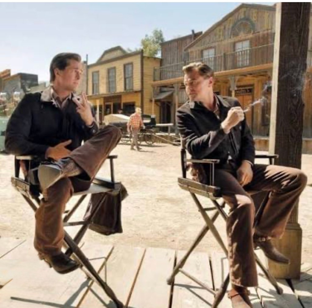 Two men sitting on directors' chairs in a Western film set, with wooden buildings in the background.