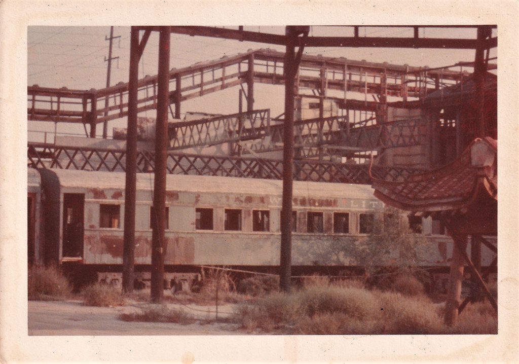 An old, weathered train car partially obscured by overgrown vegetation and structural beams in an industrial area.