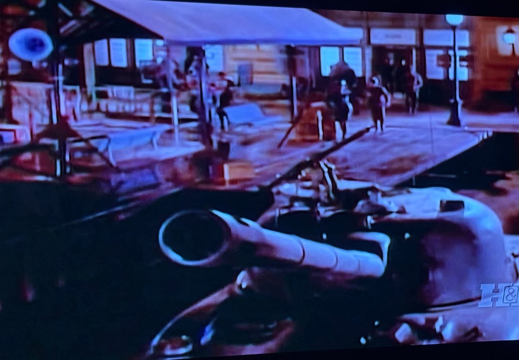 A scene depicting a military tank in the foreground with a town square in the background, featuring people walking and an old building with a canopy.