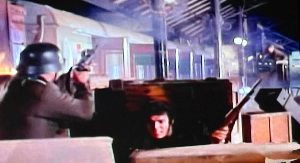 A tense scene depicting a soldier in a helmet aiming a gun from behind wooden crates, while another figure crouches nearby, suggesting a military confrontation.