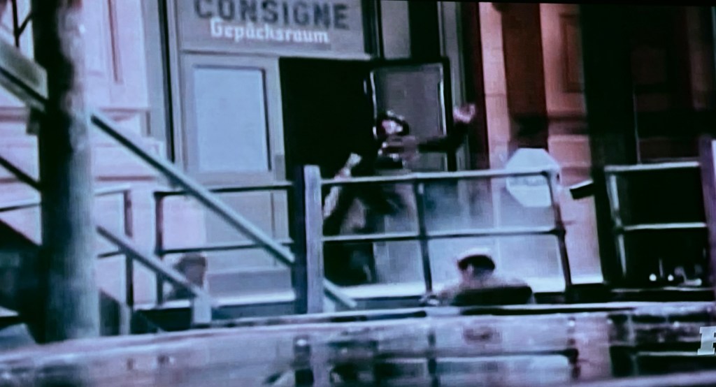 A black-and-white scene depicting a person gesturing from a doorway labeled 'Consigne Gepäckraum', with stairs leading down and a person visible below.