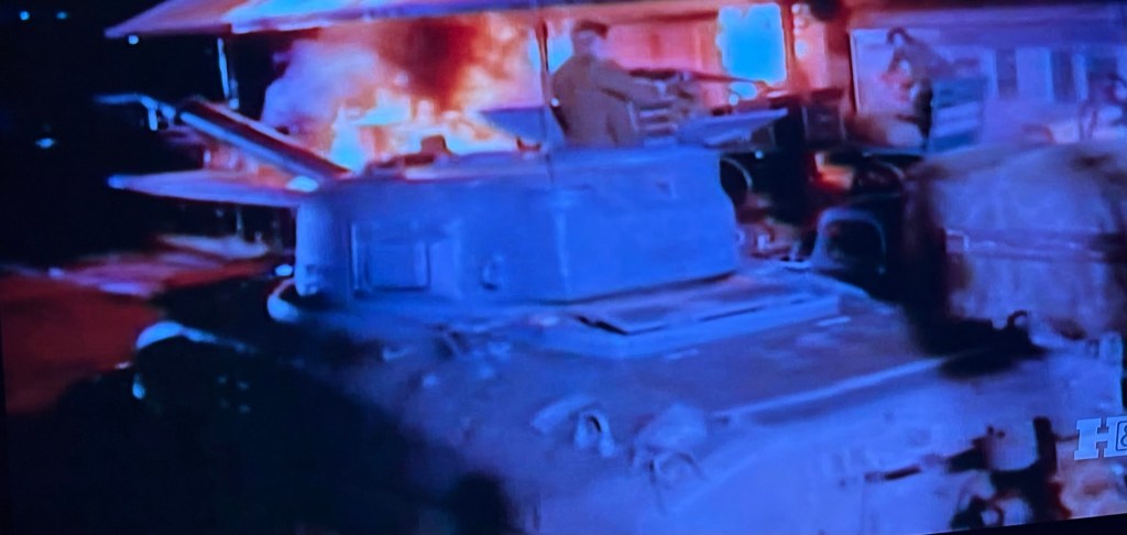 A military tank is parked near a burning structure, with soldiers present nearby.