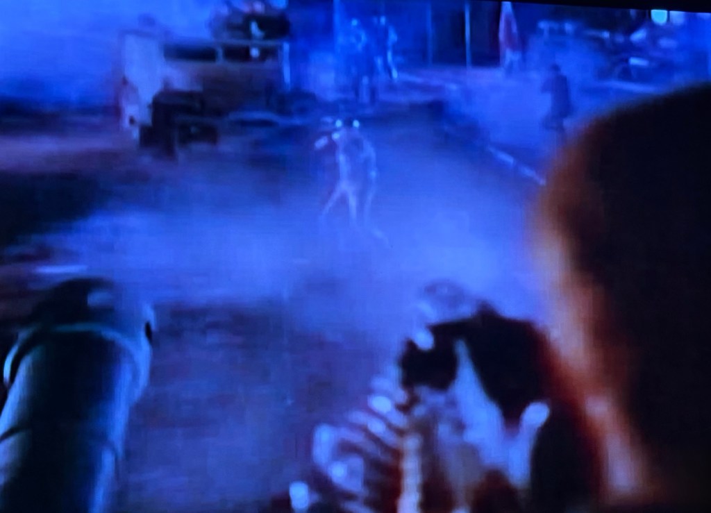 A dark, foggy scene with a figure illuminated in the distance, suggesting tension or conflict, set in an outdoor environment with military equipment.