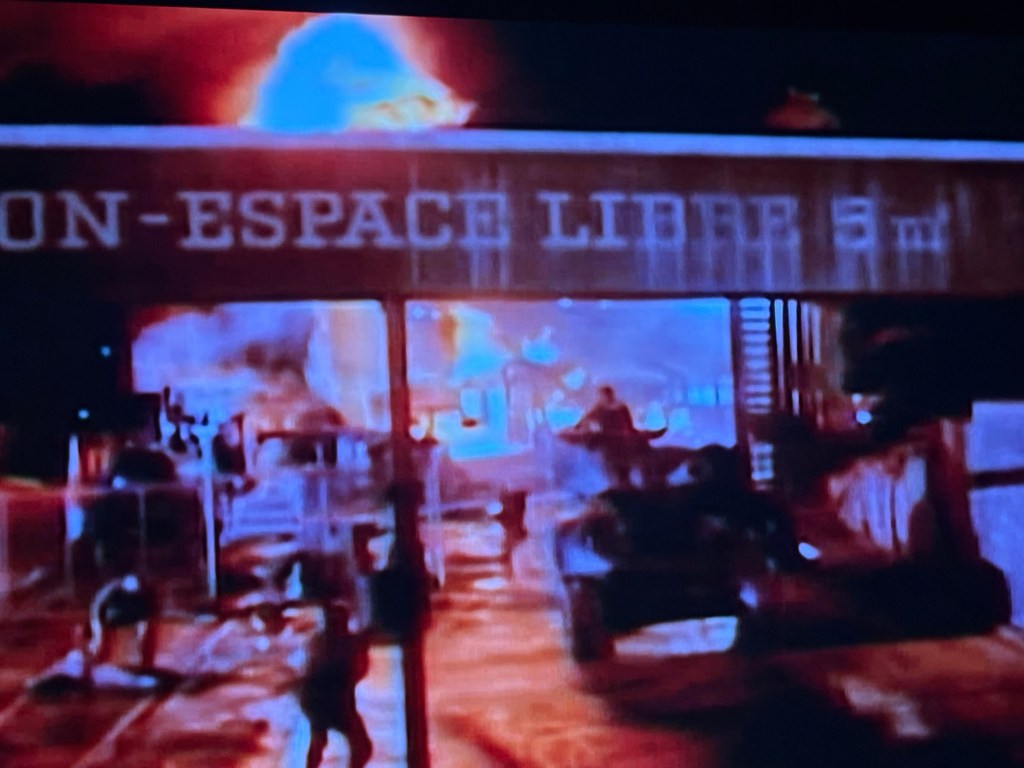 A chaotic scene with flames and smoke in an urban setting, featuring people and vehicles, under a sign that reads "ON-ESPACE LIBRE 5 m".