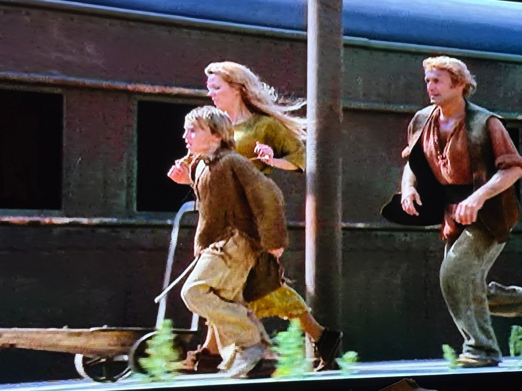 A woman and two children running alongside a train track, with a wooden cart being pushed by one of the children.