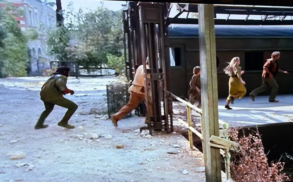 A group of people running away from a structure, with a train in the background and trees and buildings visible.