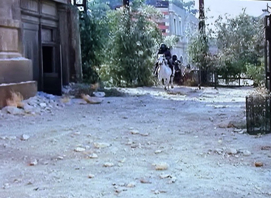 A dirt road with scattered rocks, leading past a partially visible building doorway, with two riders on horseback approaching, surrounded by greenery.