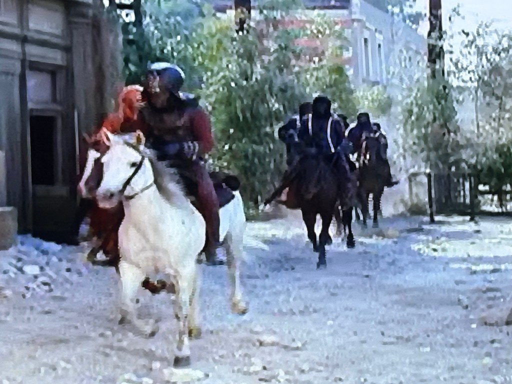 A rider on a white horse leads a group of mounted figures through a dusty street lined with greenery and buildings.