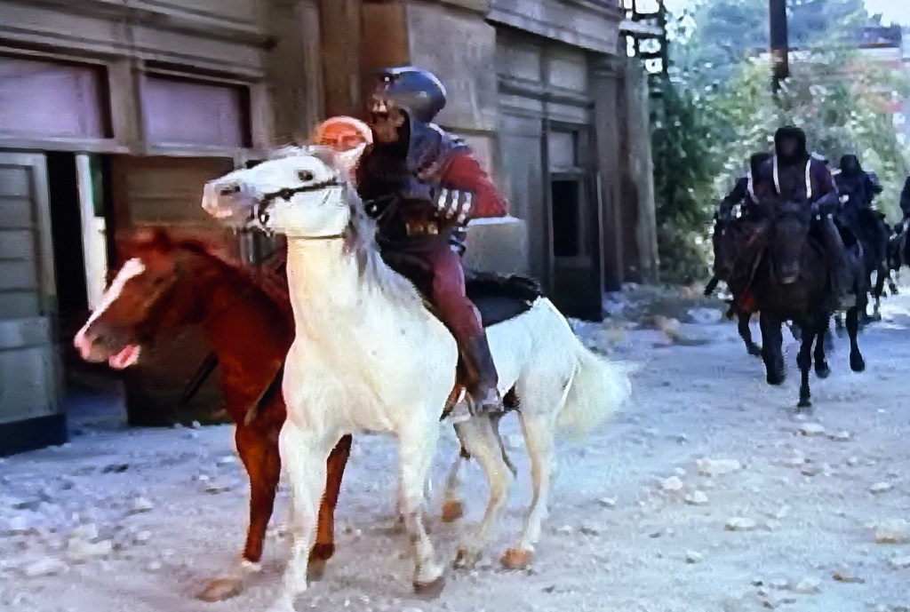 A rider on a white horse and another on a brown horse are galloping through a partially deserted street, with several riders on black horses visible in the background.