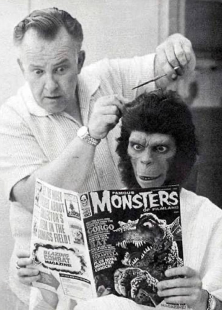 A man styles the hair of a person in a gorilla costume who is reading a magazine titled 'Famous Monsters of Filmland.' The scene captures a humorous interaction between the hairdresser and the costumed individual.