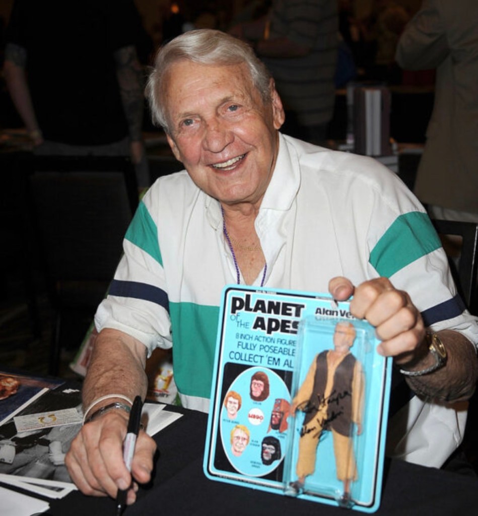 An elderly man with white hair smiling and holding an action figure package from 'Planet of the Apes', signed by him, while sitting at a table.