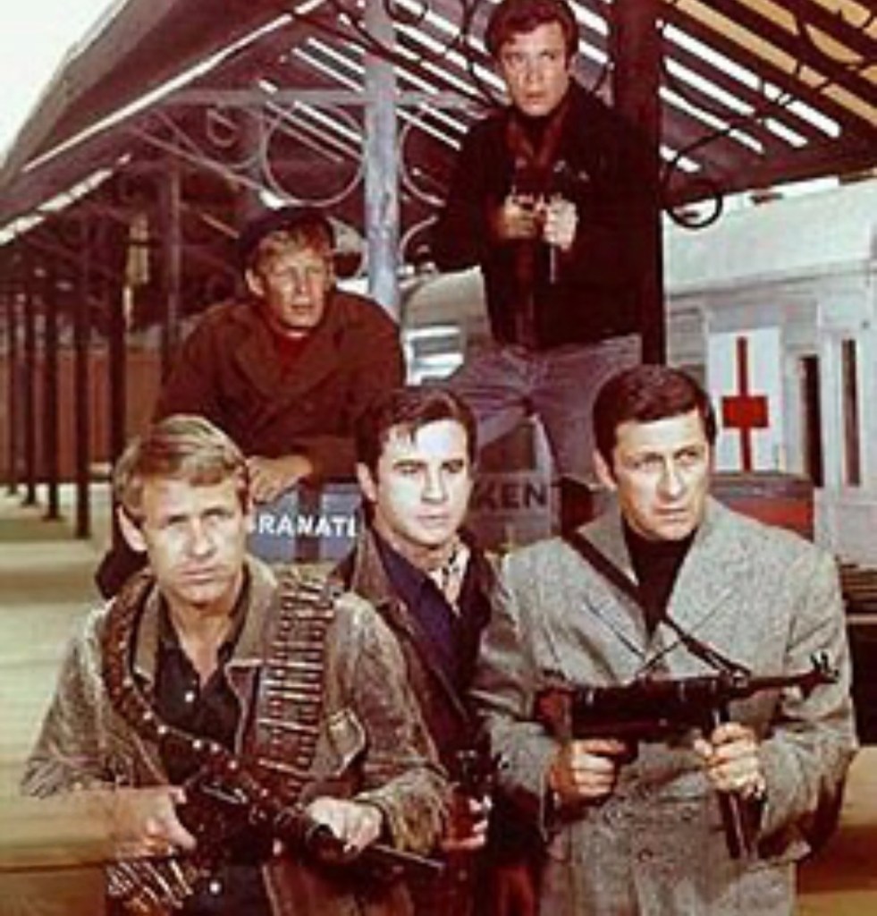 A group of five men posed in a warehouse or train station, some holding guns and looking serious. The background features train tracks and industrial elements.
