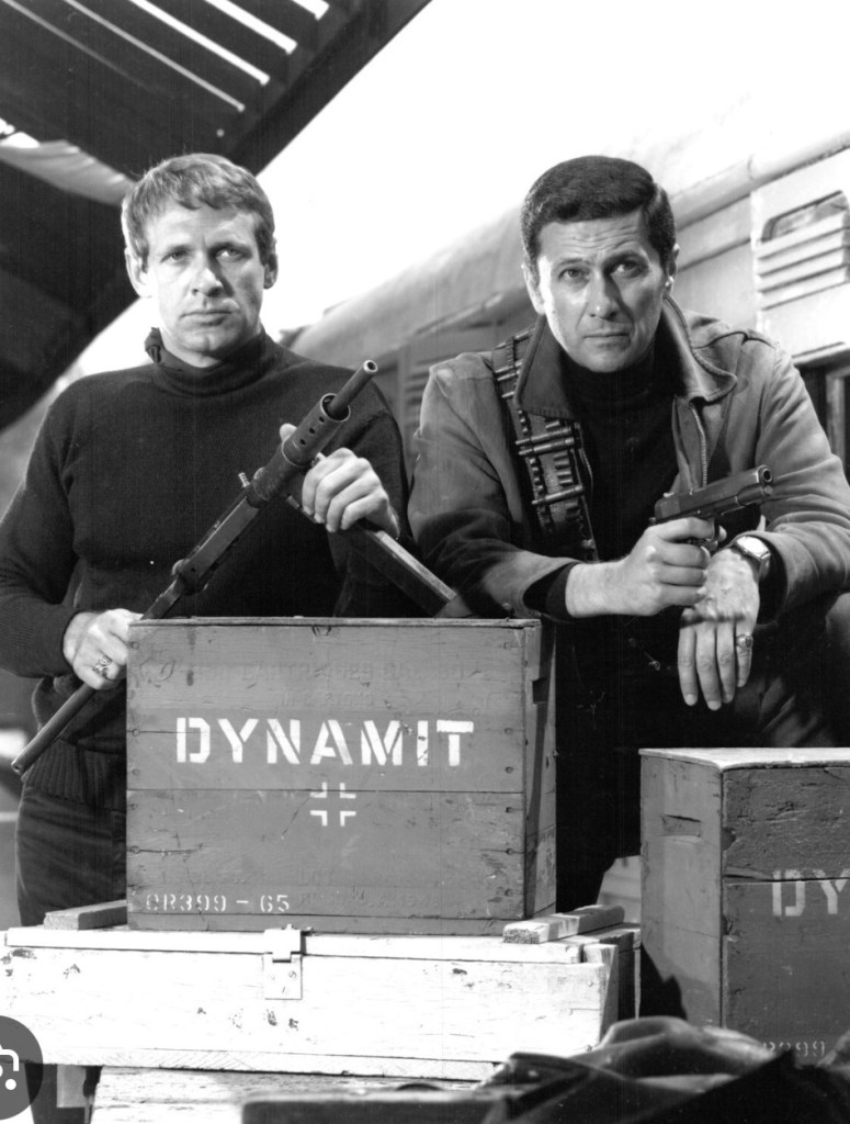 Two men in black and white, posing with firearms, stand beside wooden crates labeled 'DYNAMIT.' The setting appears to be a train station.