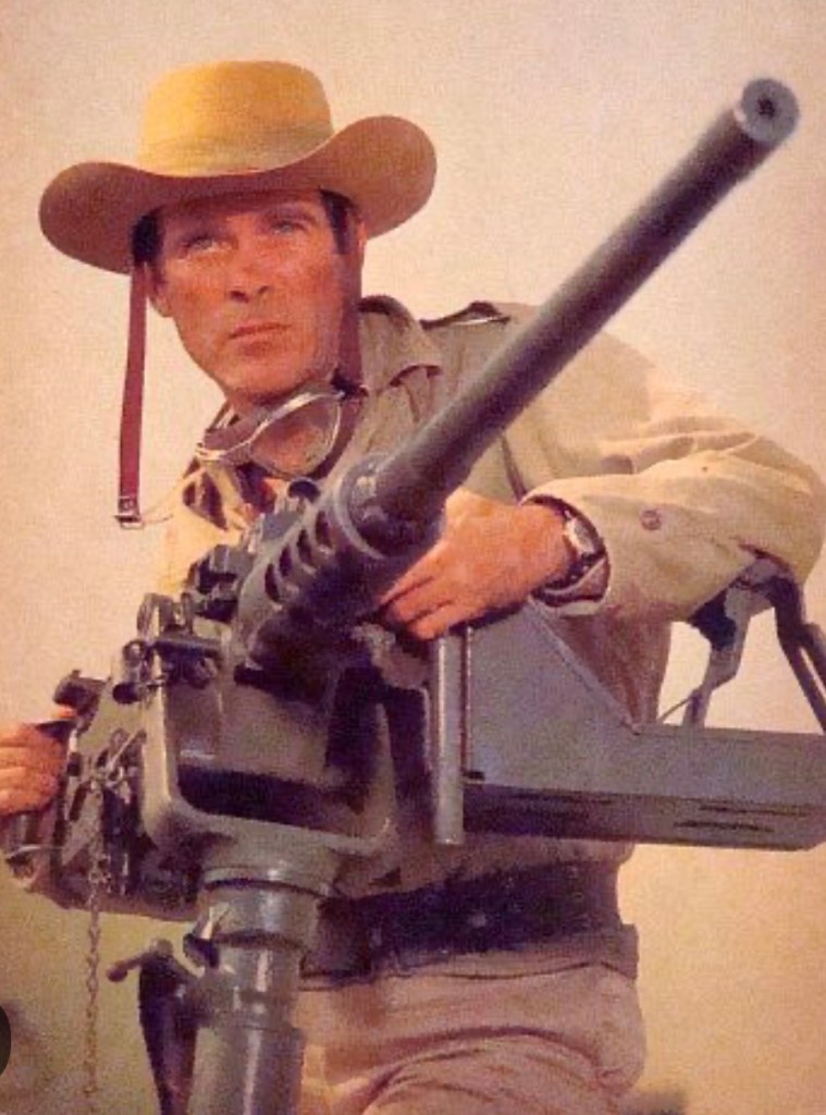 A man wearing a wide-brimmed hat holds a large machine gun, looking intently at the camera.