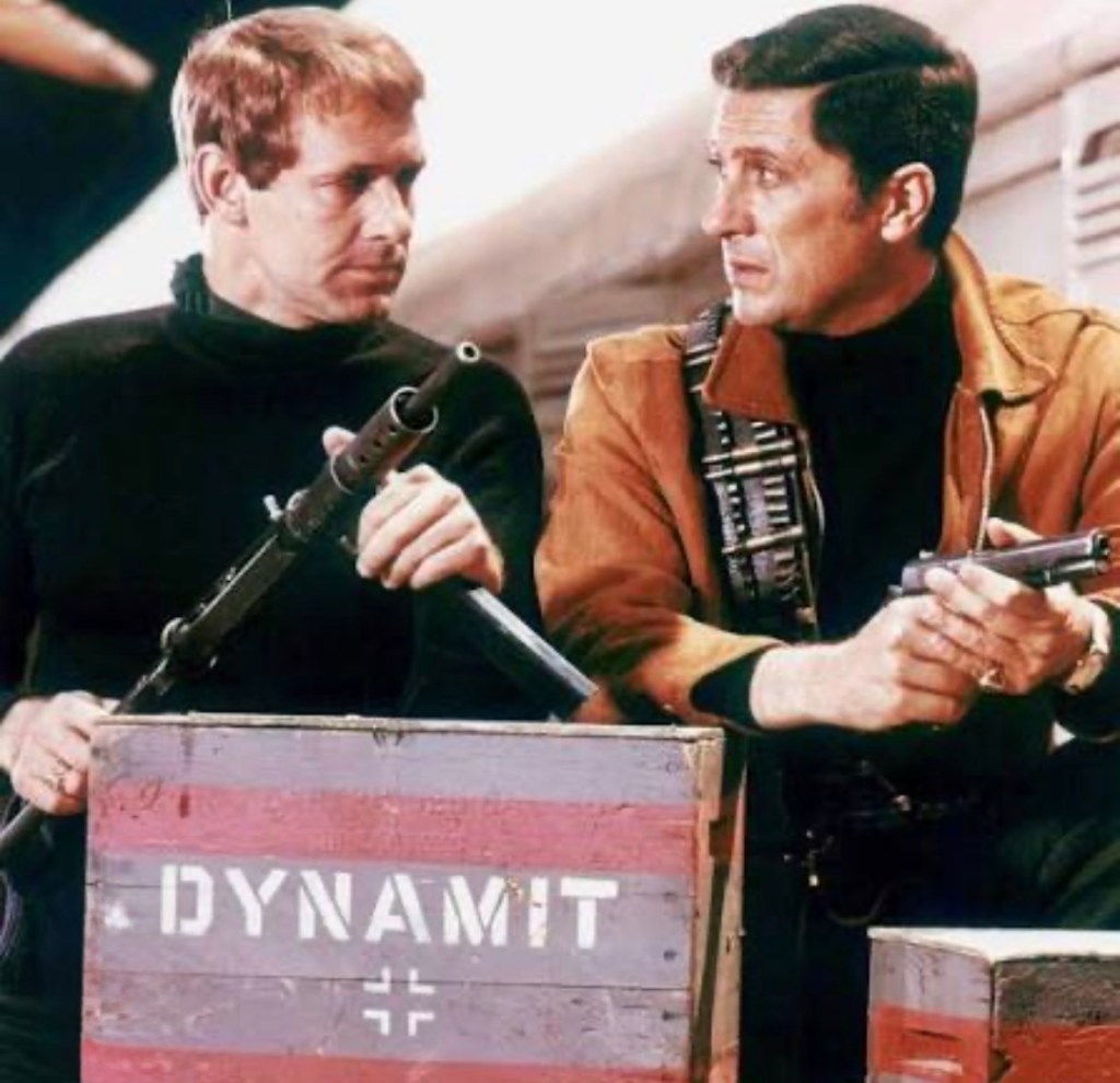 Two men holding weapons while standing next to wooden crates marked 'DYNAMIT' in a dramatic setting.