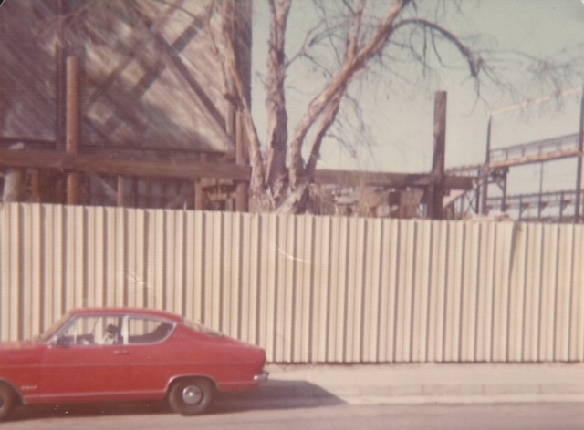 A red car parked beside a corrugated metal fence, with a tree and building structure visible in the background.