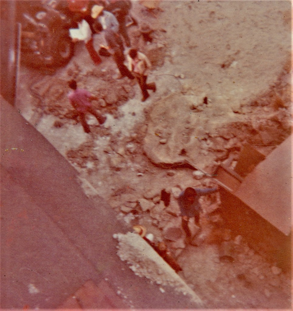 A top-down view of a construction site with several people walking over uneven ground, surrounded by rubble and debris.