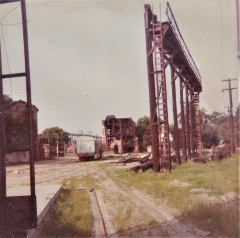 Abandoned industrial site with a partially demolished building in the background and rusty metal structures in the foreground, overgrown with grass.