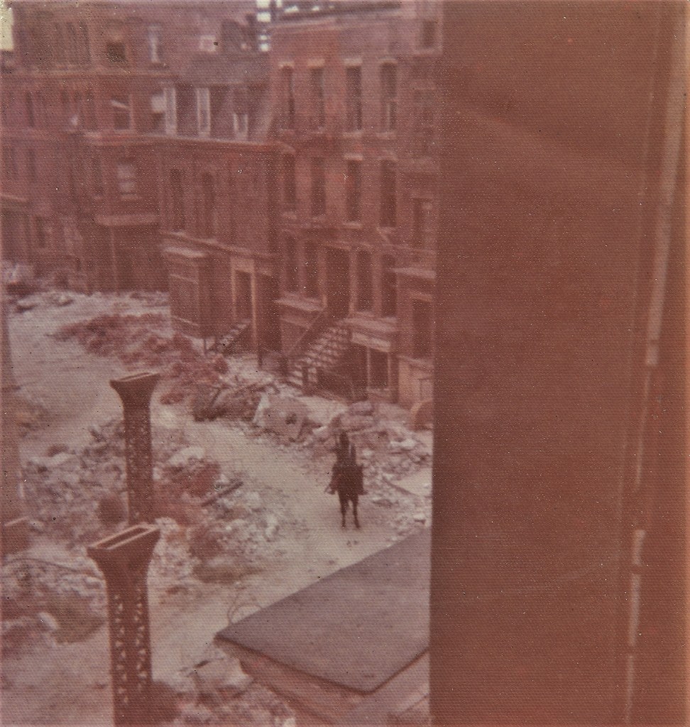 A view of a dilapidated street with rubble, featuring old brick buildings with fire escapes and a solitary figure on horseback walking through the debris.