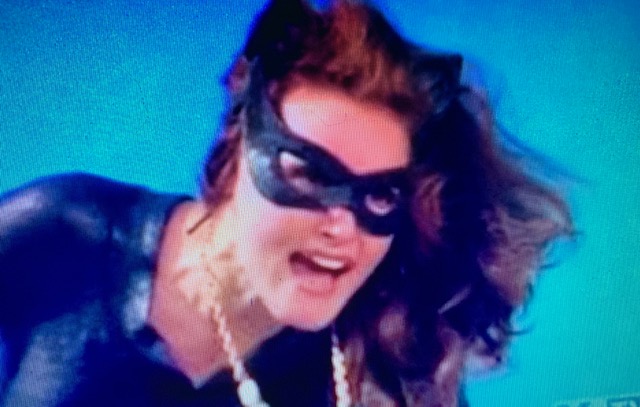 Woman wearing a black cat mask and costume, with flowing hair, expressing a fierce appearance against a blue background.