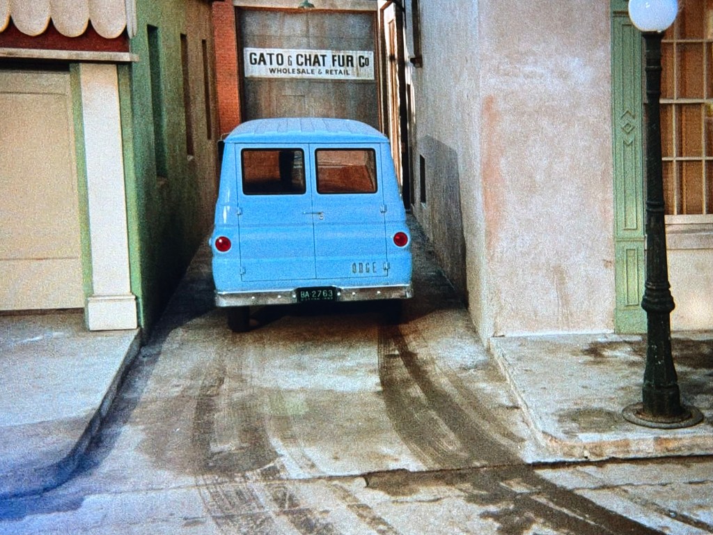 A light blue van parked in a narrow alleyway, partially obstructed by walls on either side. A sign reading 'GATO & CHAT FUR CO. WHOLESALE & RETAIL' is visible in the background. The ground shows tire tracks leading into the alley.