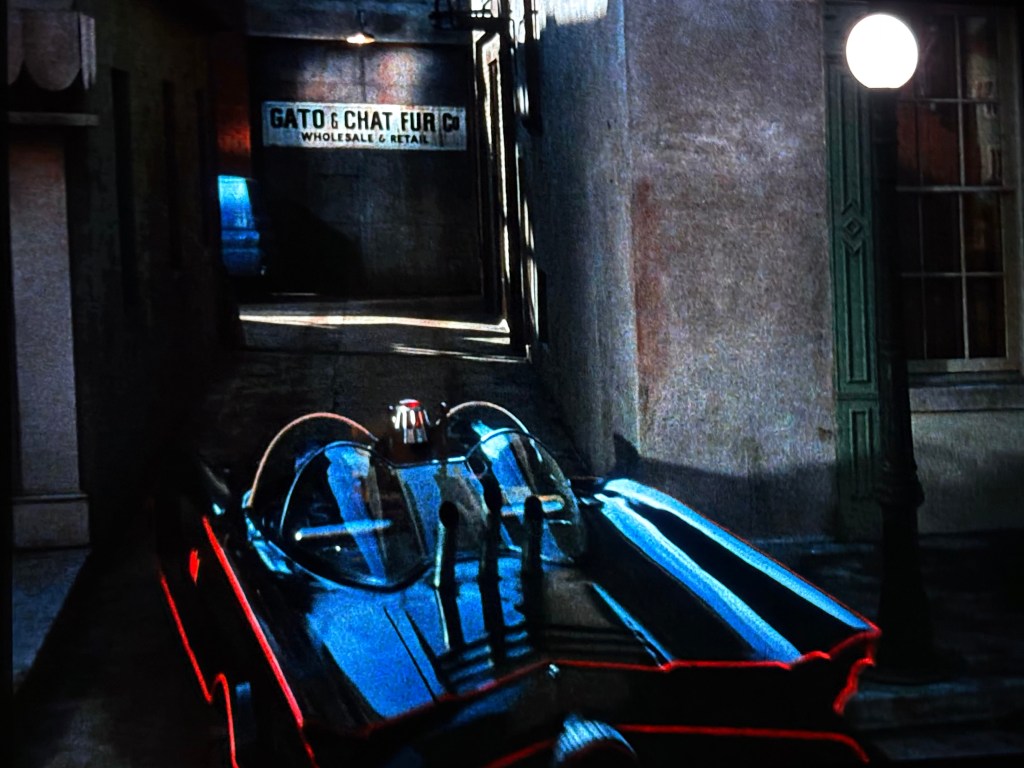 A street scene featuring a sleek, black car with bright red accents parked in an alleyway. A sign on the wall reads 'GATO & CHAT FUR CO. WHOLESALE & RETAIL'.