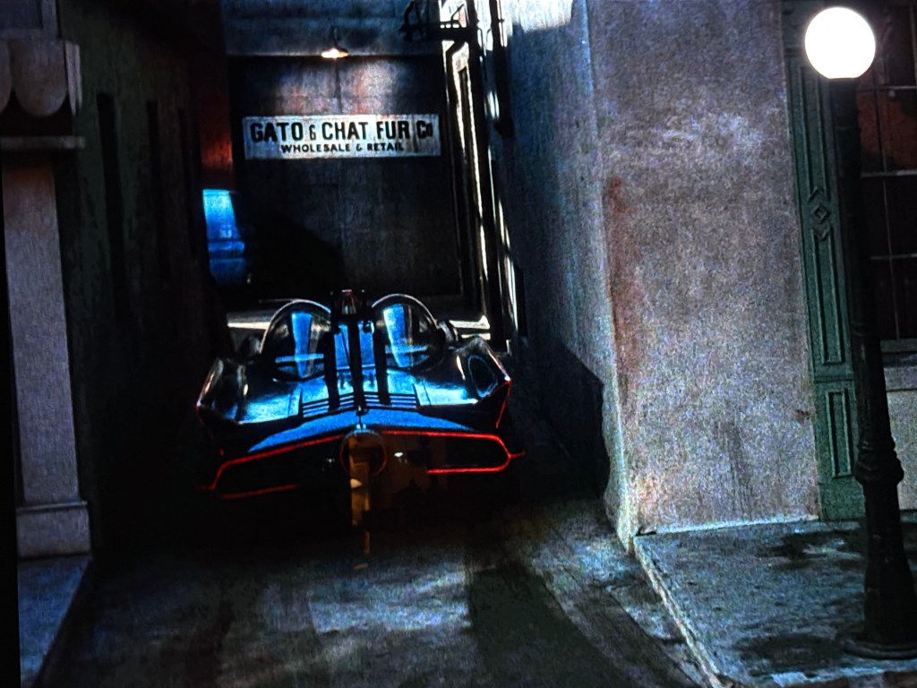 A sleek blue and black futuristic car parked in a narrow alleyway, with a sign reading 'Gato & Chat Fur Wholesale & Retail' visible in the background.