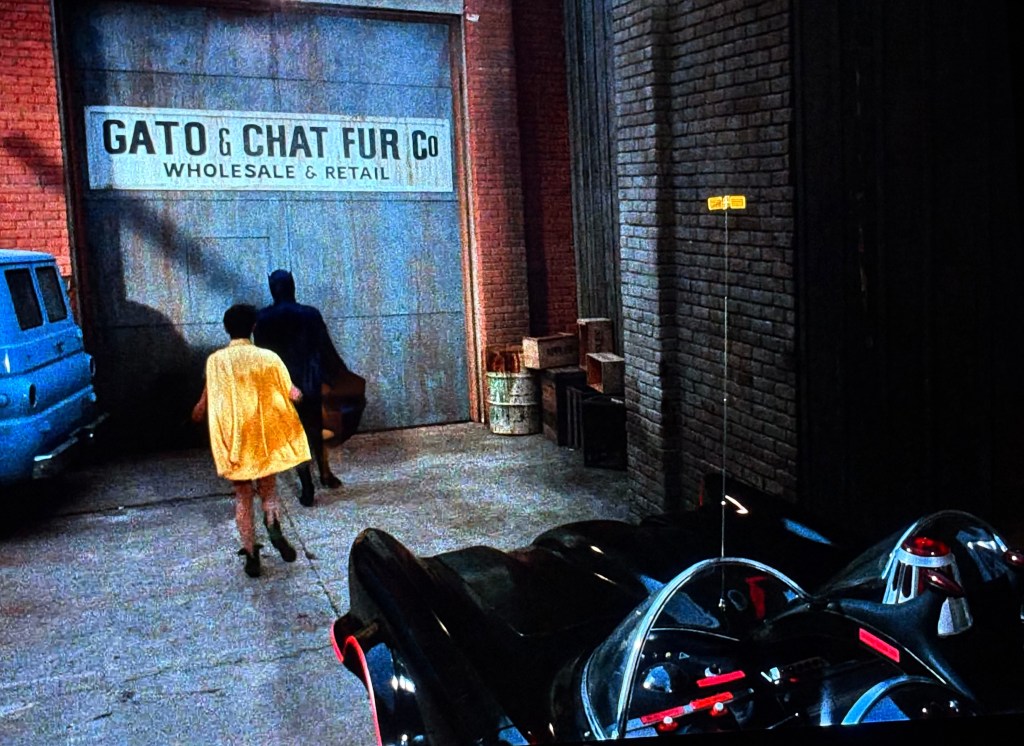 A yellow-coated figure and a dark-clad figure walk towards a warehouse marked 'GATO & CHAT FUR Co' on a brick wall. An iconic black vehicle is parked in the foreground.