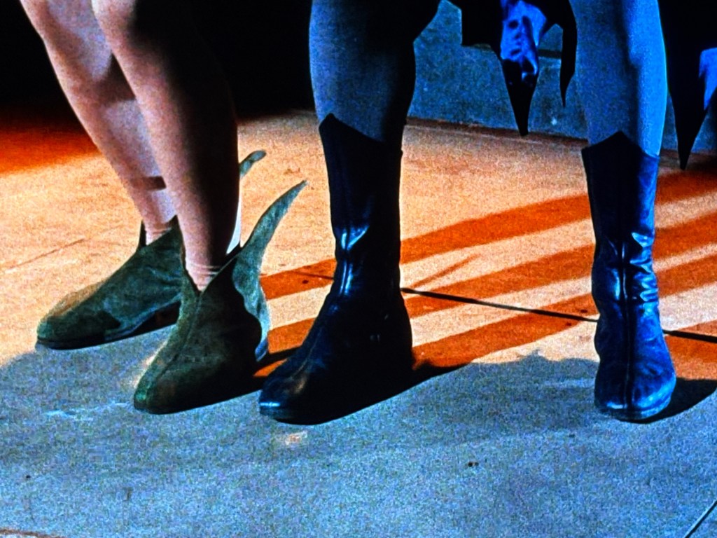 A close-up shot of two pairs of shoes, one green suede boot with a unique design and one black leather boot, alongside a pair of bare legs. The background features a dimly lit stage.