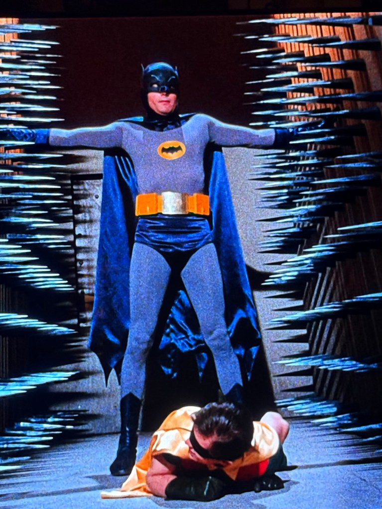 Batman stands heroically with his arms outstretched in a dramatic pose, while Robin kneels on the ground in front of him, suggesting a tense scene.