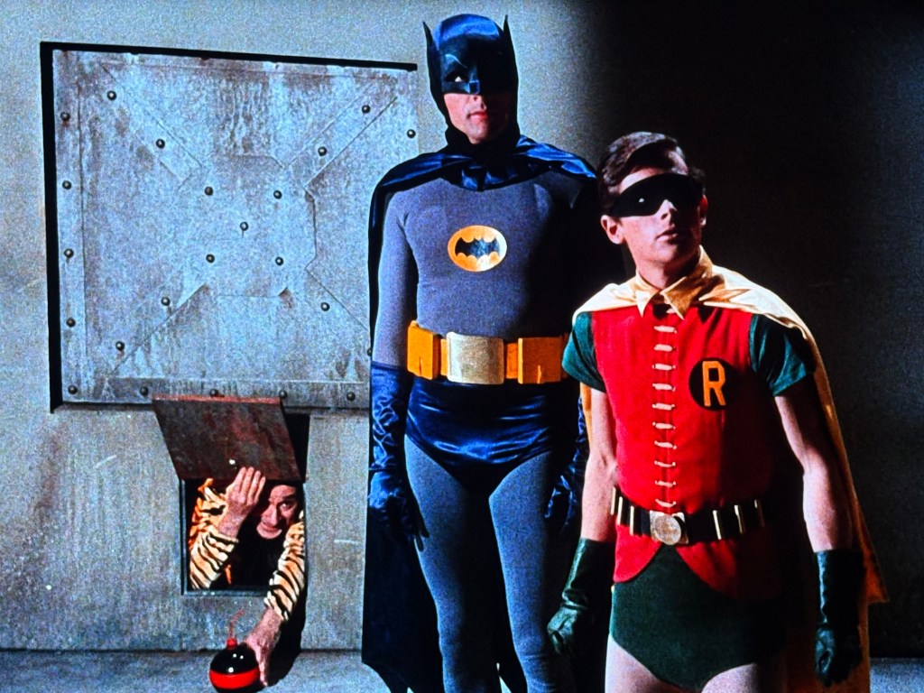 Batman and Robin stand in a dimly lit room, with Batman in a gray suit and Robin in a red and green costume. A villain in stripes can be seen peeking out from a small opening in the background.