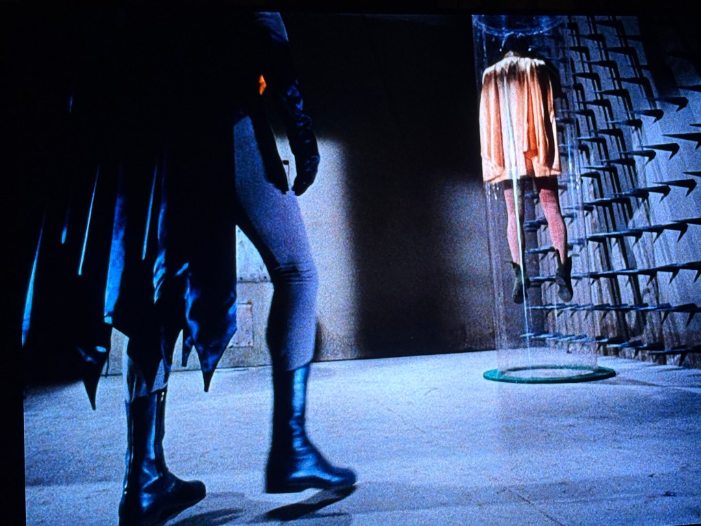 A figure in a dark cape approaches a transparent containment chamber holding a suspended character in an orange outfit, set against a stark industrial background.