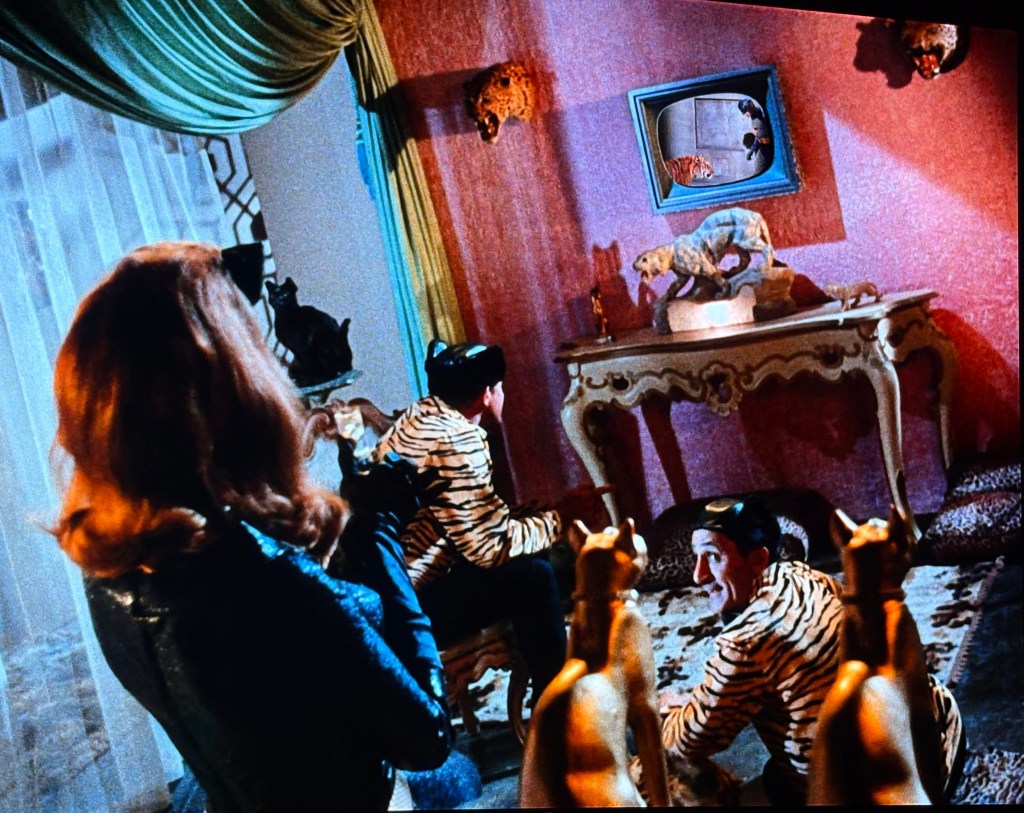 A room featuring a woman in a black outfit and two men dressed in tiger-striped suits, sitting on a rug while watching a television displaying a tiger. Decorative animal sculptures and a vintage style table are also visible.