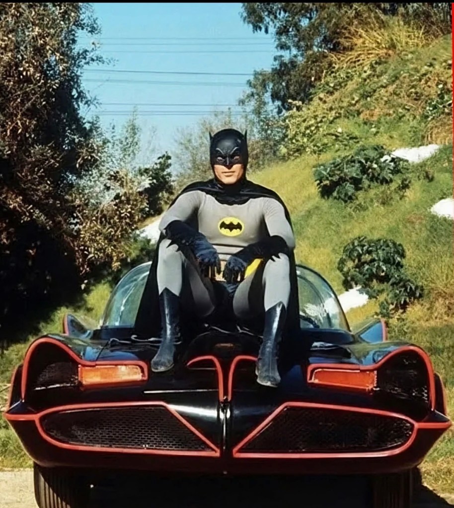 Batman in a classic gray and black costume sitting on the hood of a retro Batmobile, surrounded by greenery.
