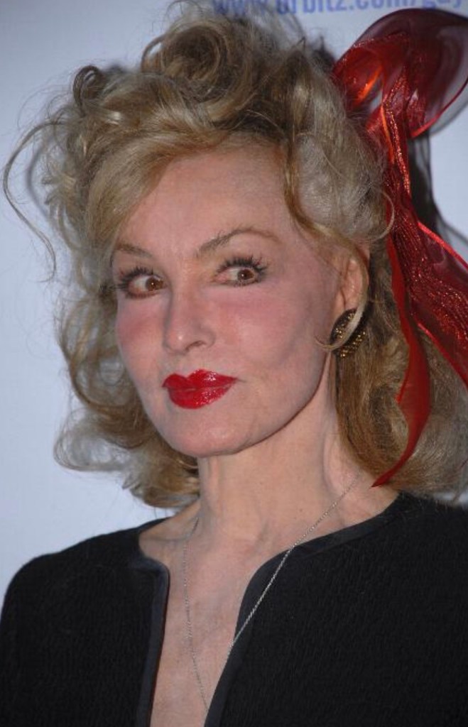 A woman with wavy blonde hair and a red ribbon, wearing bold red lipstick and a black outfit, posing for the camera.