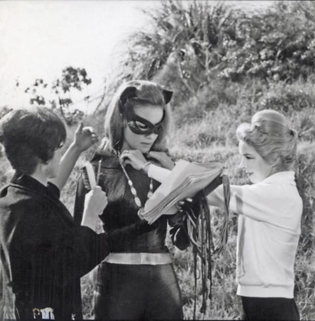 Behind-the-scenes image of a woman dressed as a cat-themed character being styled by two assistants, surrounded by greenery.