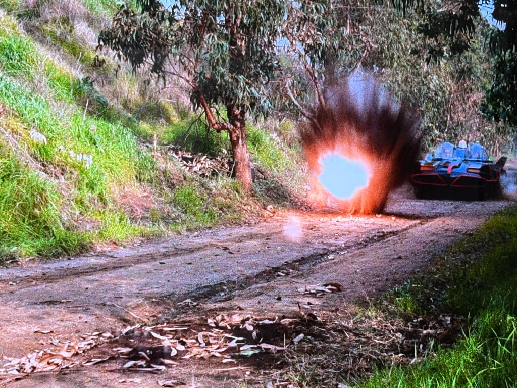 An explosion occurs on a dirt road surrounded by greenery, while a car speeds by.