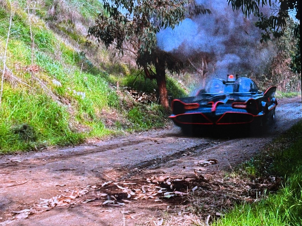 A classic black car with red accents, resembling a Batmobile, driving down a dirt road surrounded by lush greenery and smoke coming from the vehicle.