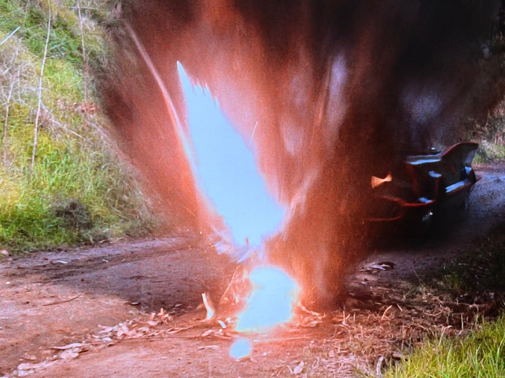 An explosion occurring on a dirt road, with bright flames and smoke rising, while a car is visible in the background.