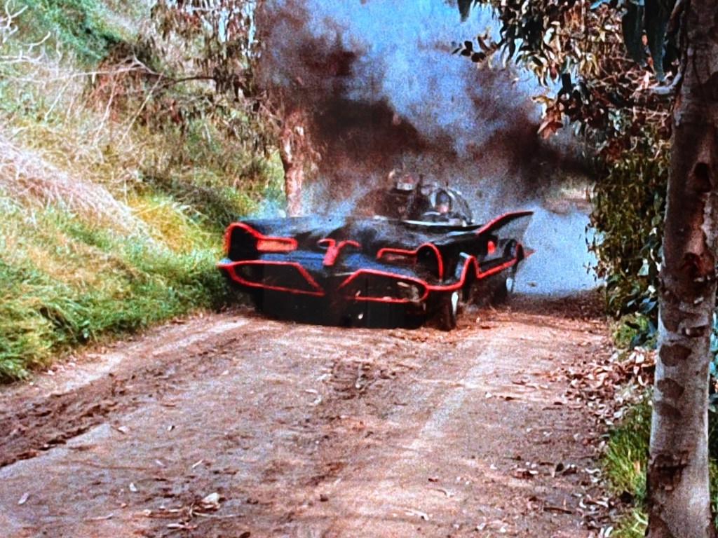 A classic black and red batmobile driving down a dirt path, emitting smoke in a wooded area.