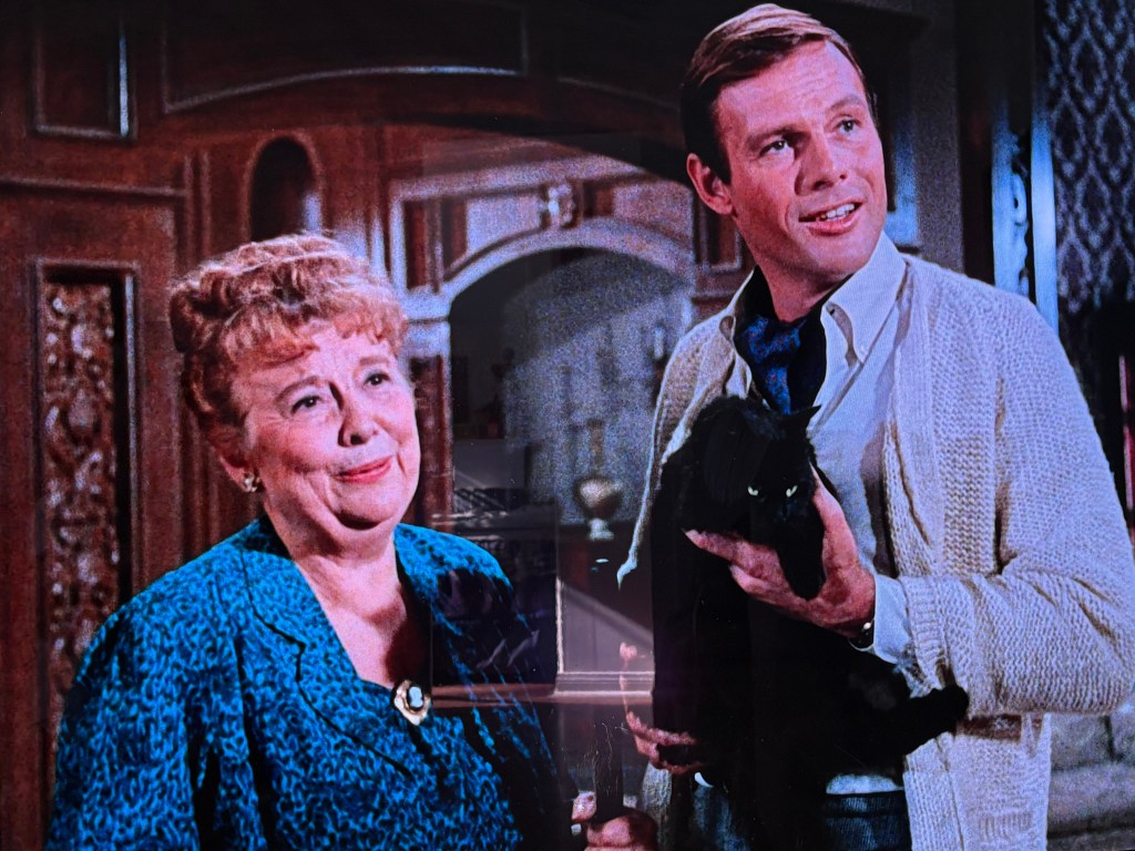 A man holding a black cat smiles while standing next to a woman with curly hair, who looks pleased; both are in a warmly decorated interior.